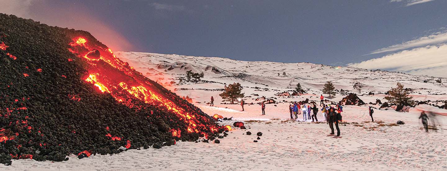 People observe glowing red lava flowing down a snowy volcanic slope