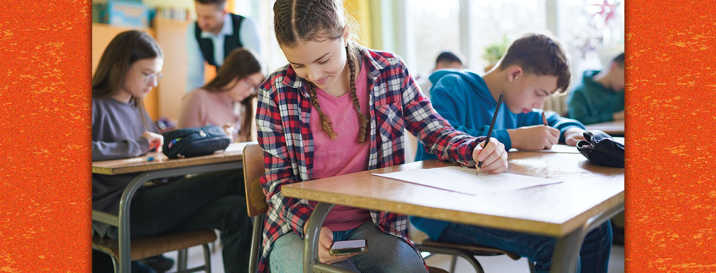 A girl writes on paper at a desk while she holds a phone, surrounded by classmates
