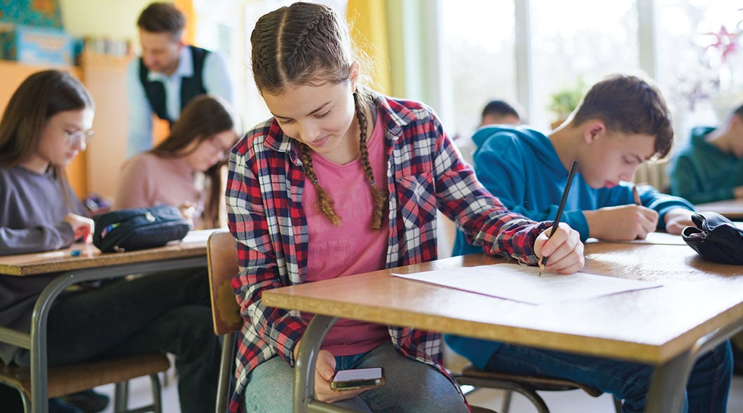 A girl writes on paper at a desk while holding a phone, surrounded by students in a classroom
