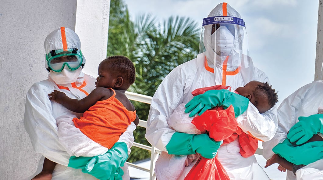 Two healthcare workers in full protective gear and face shields each hold a young child