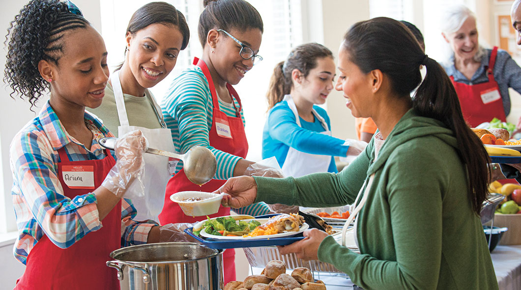 A group of volunteers in red aprons serve food to a woman at a community meal event