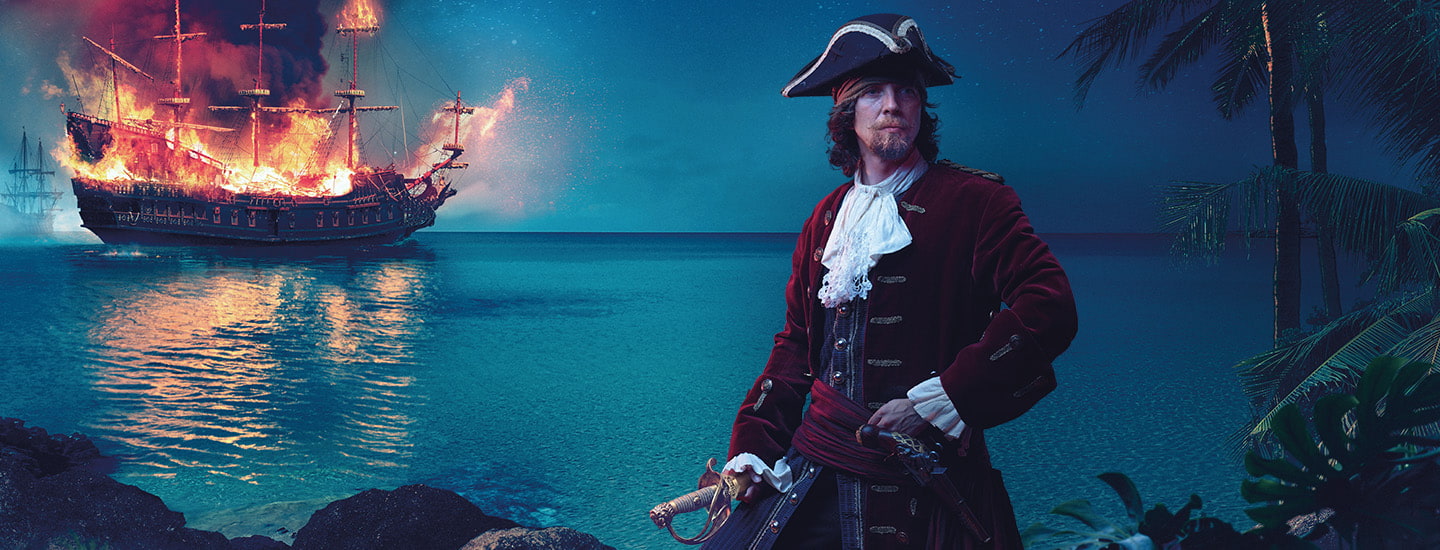 A pirate holds a sword on a shore while a tall ship burns on the water behind him