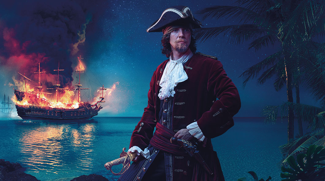A pirate holds a sword near a tropical shore while a tall ship burns on the water behind him
