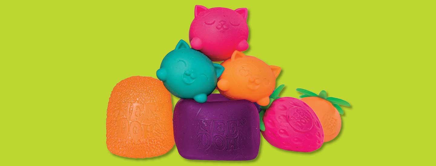 Three cat-faced squishy toys sit atop an orange gumdrop, purple bar, strawberry, & pineapple