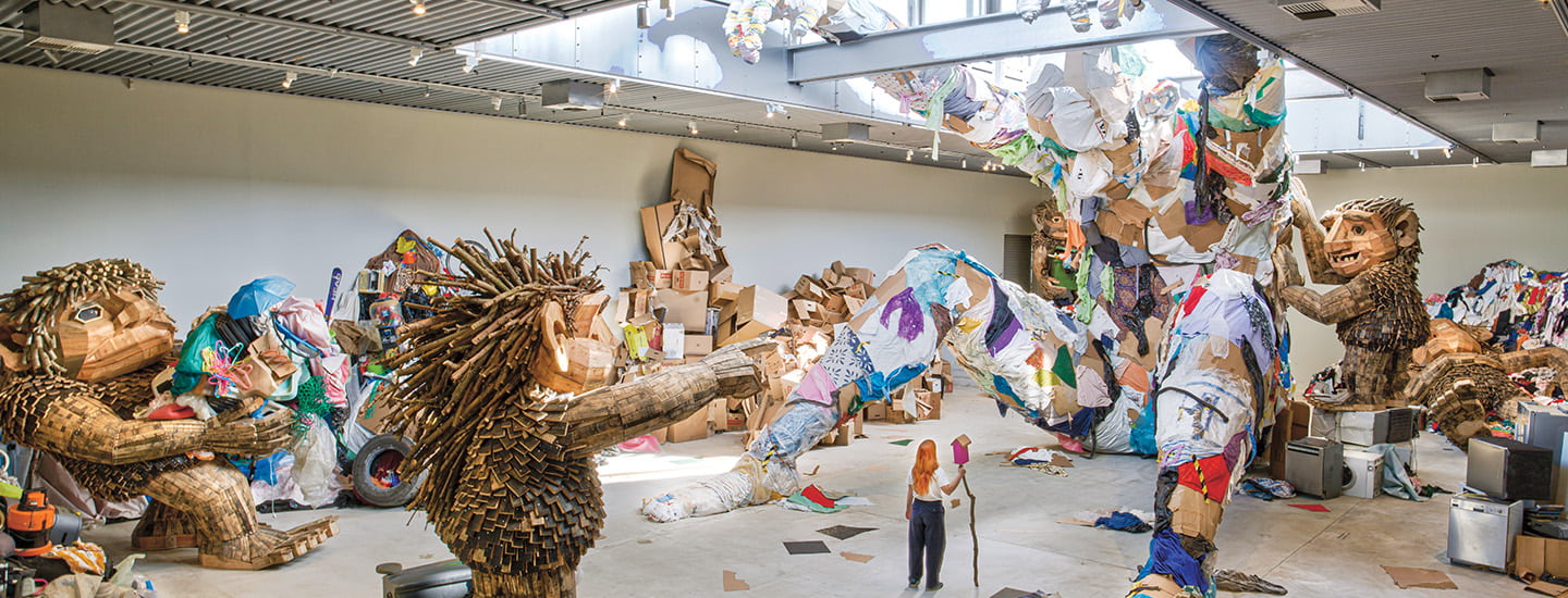 A gallery installation with massive sculptures made from recycled materials towers over a small figure below